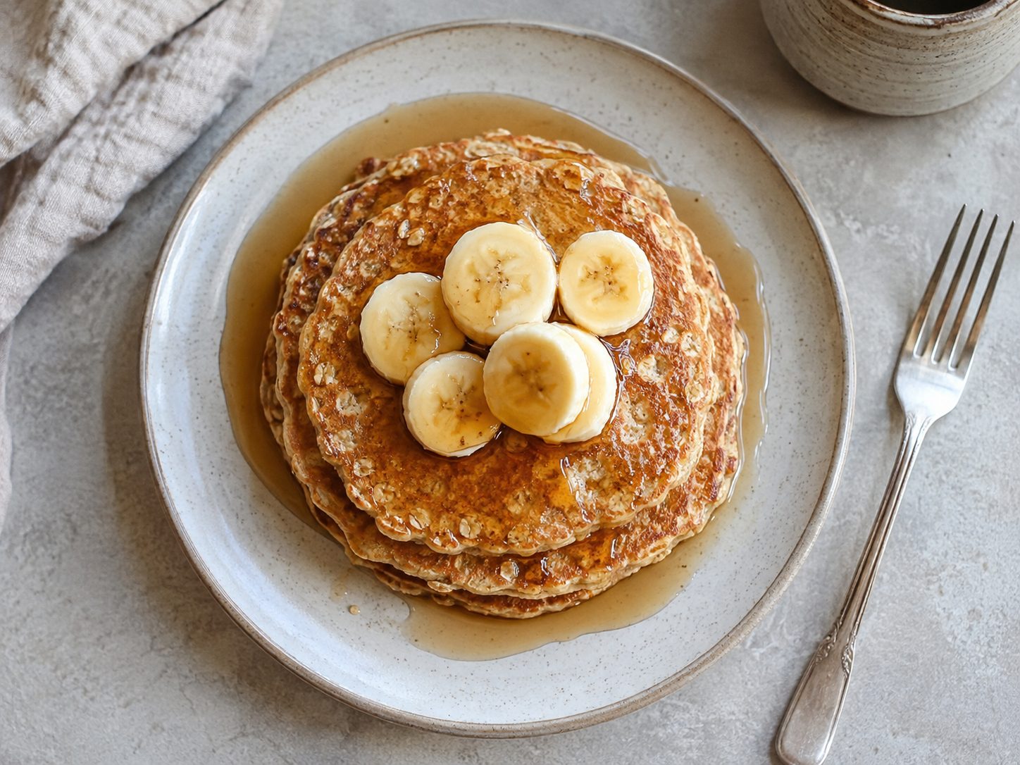 3-Ingredient Banana Oat Pancakes for a Fast, Flourless Breakfast 3 Overhead view of banana oat pancakes stacked on a plate with syrup and a fork