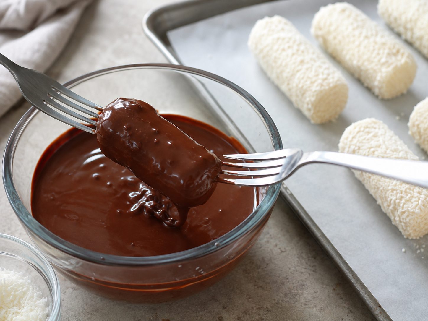 3-Ingredient Healthy Mounds Bars: A No-Bake Coconut Candy You Shape by Hand 2 Forks dipping a chilled coconut log into a bowl of melted chocolate for Mounds bars